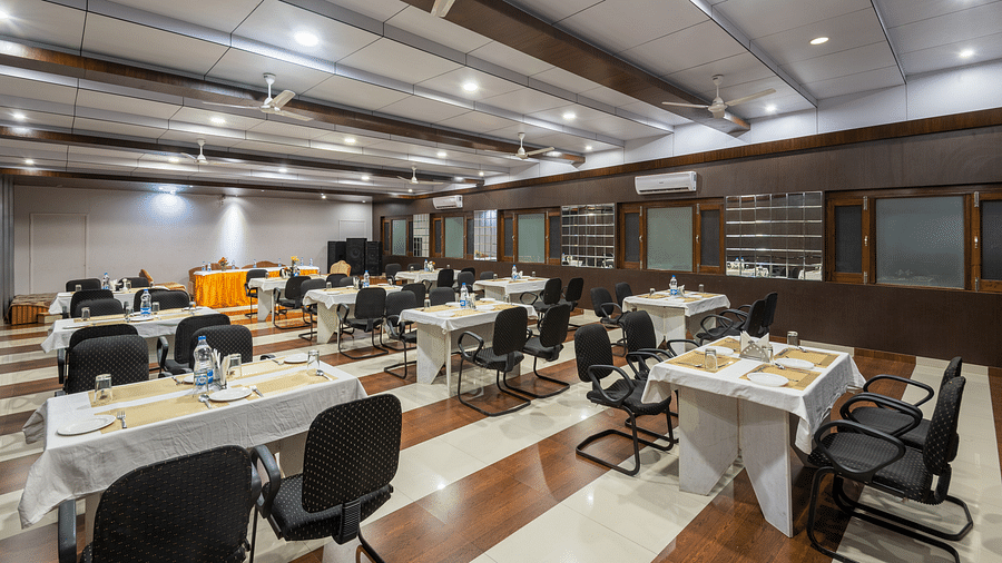 A well-lit conference room with rows of tables and chairs, featuring a projector screen and a fan - DLS Meghavan Resort